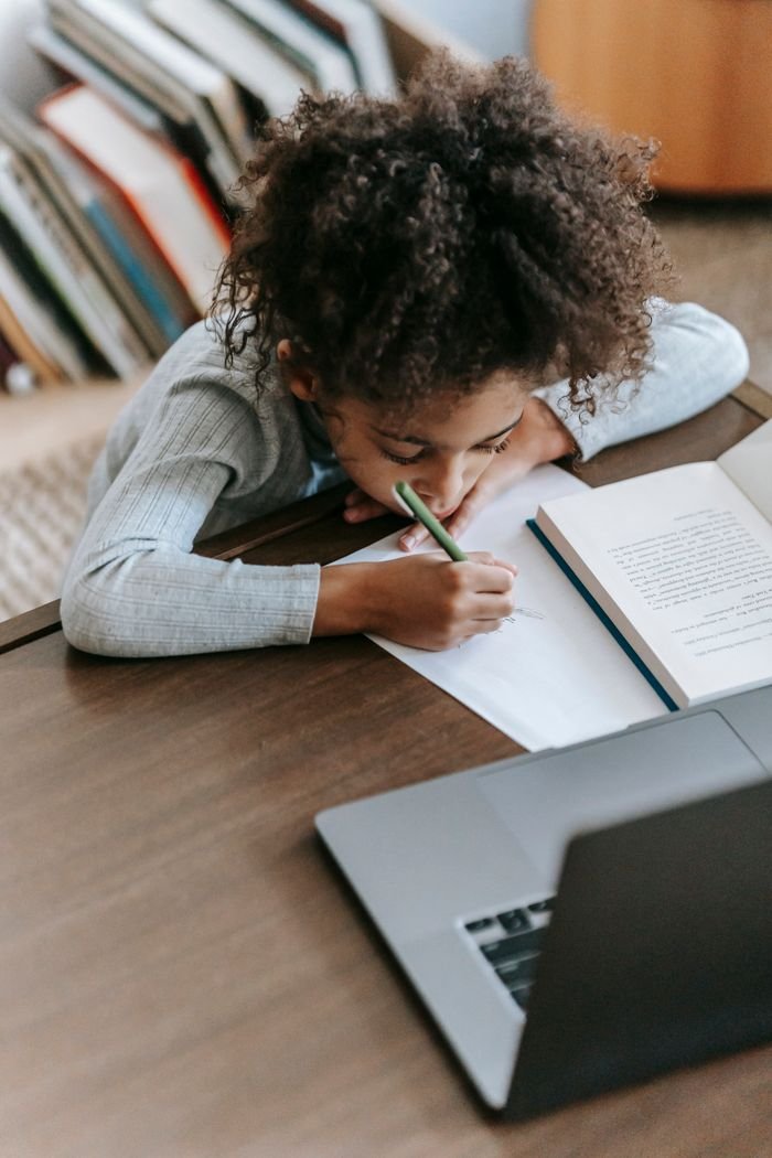 about-us-02 High angle of diligent calm African American schoolchild with curly hair in casual clothes taking notes in copybook sitting at table with netbook during exam preparation at home