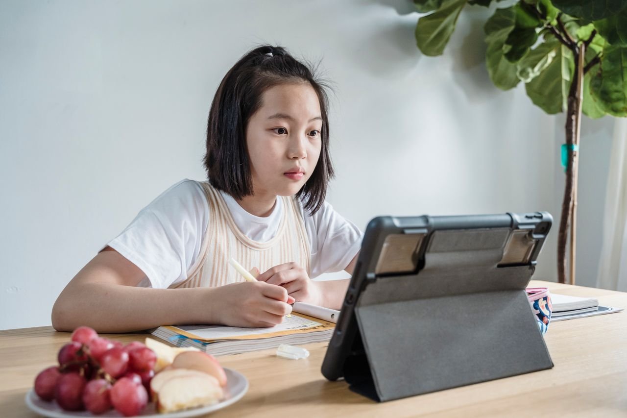 about-us-01 Young girl engaged in online learning using a tablet, seated at a table with snacks indoors.
